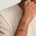 Man wearing a gold bracelet with a blue charm on a plain background