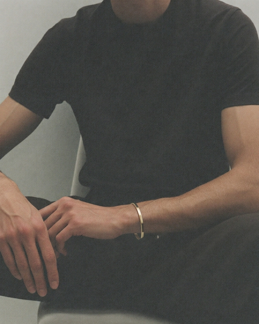 A man wearing a simple gold bangle bracelet on his wrist, dressed in a black t-shirt and black trousers while sitting.