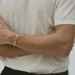 A close-up of a person wearing a slim silver bangle bracelet on their left wrist, paired with a cream-colored t-shirt and black trousers.