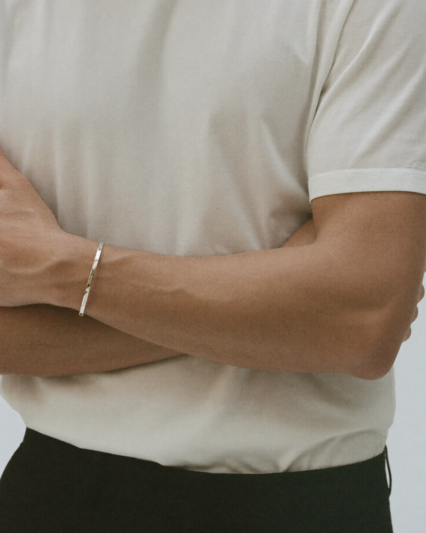 A close-up of a person wearing a slim silver bangle bracelet on their left wrist, paired with a cream-colored t-shirt and black trousers.